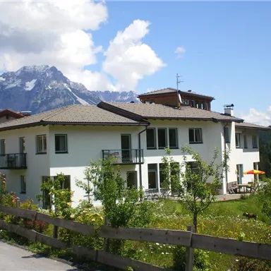 A modern house with a balcony, surrounded by green landscape and flowers. In the background, the mountains are visible under a blue sky with some clouds.