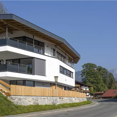 A modern house with a large terrace and panoramic windows. In the background, mountains and traditional buildings can be seen.