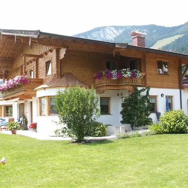 A beautiful wooden house with a balcony and colorful flowers. It is located in a green environment, surrounded by mountains.