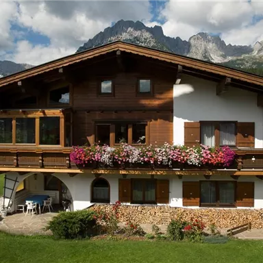 A traditional wooden house with colorful flowers on the balcony. In the background, majestic mountains and a clear sky can be seen.