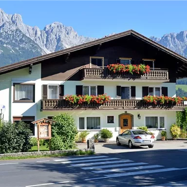 Ein traditionelles alpenländisches Haus mit Blumenkästen und einem klaren blauen Himmel. Im Hintergrund sind beeindruckende Berggipfel sichtbar.