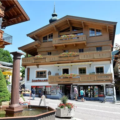 Ein charmantes Gebäude im alpinen Stil mit Balkonen und bunten Blumen. Im Vordergrund befindet sich ein Brunnen und Besucher erkunden die Umgebung.