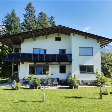 Ein modernes Haus mit Solarpanelen und einem gepflegten Garten. Umgeben von Bäumen und Bergen, bietet es eine schöne Aussicht.