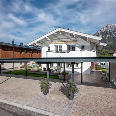 Ein modernes Haus mit einem großzügigen Carport und einem gepflegten Garten. Im Hintergrund sind beeindruckende Berge zu sehen und der Himmel ist leicht bewölkt.