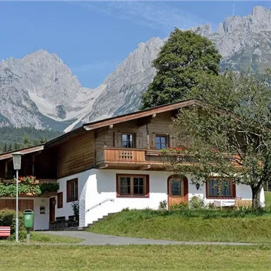 Ein traditionelles Haus in den Bergen mit einer grünen Wiese und Bäumen. Im Hintergrund sind majestätische, felsige Gipfel zu sehen.