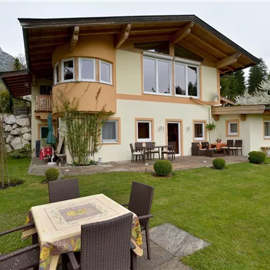 Ein schönes Haus mit einem großen Garten und Terrase. Im Hintergrund sind Bäume und Berge zu sehen.