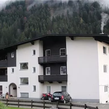 Ein modernes Gebäude in den Bergen, umgeben von grüner Natur. Rauch steigt von einem Schornstein auf, während Wolken die Gipfel umhüllen.