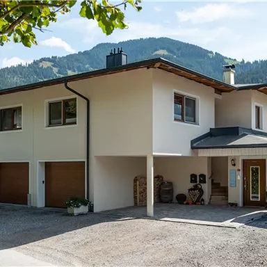 A modern house in a rural setting with green mountains in the background. The façade is bright and friendly, surrounded by trees.