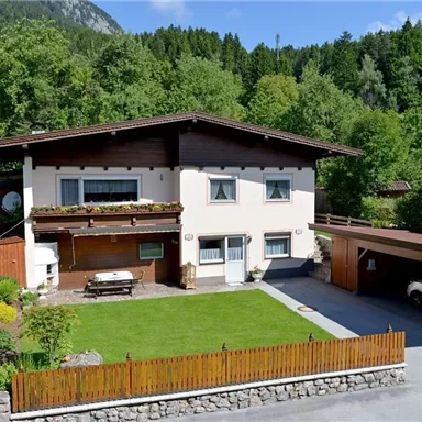 A beautiful house with a well-maintained garden and a carport. Surrounded by trees and mountains, it offers a peaceful and inviting atmosphere.