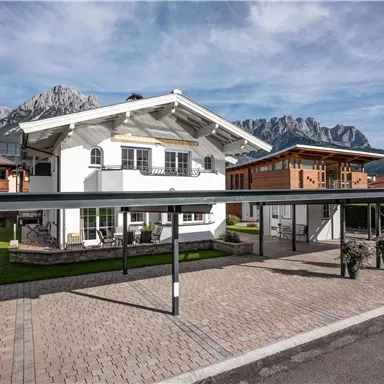 Ein modernes Haus in den Bergen, umgeben von grüner Wiese und anderen Chalets. Der Himmel ist blau und die Berglandschaft ist eindrucksvoll im Hintergrund.