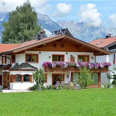 Ein charmantes Haus mit einem schönen Balkon, geschmückt mit bunten Blumen. Im Hintergrund sind beeindruckende Berge und ein blauer Himmel zu sehen.