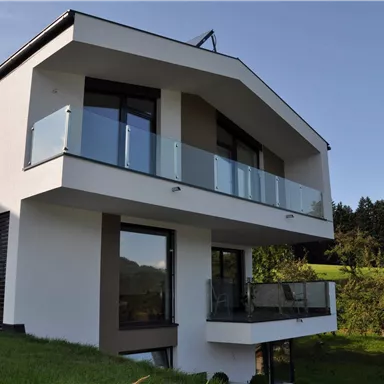 Ein modernes, zweistöckiges Haus mit großen Fenstern und einem Balkon. Im Hintergrund sind grüne Wiesen und Berge zu sehen.