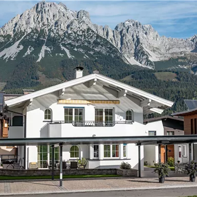 Ein schönes Haus mit einem modernen Design vor beeindruckenden Bergen. Die Umgebung ist grün und einladend.