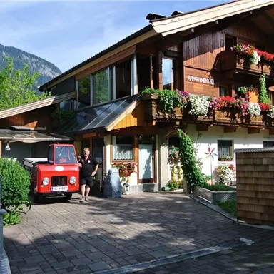 A picturesque house with flower balconies and a red car in front of it. Surrounded by an idyllic landscape and mountains in the background.