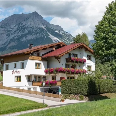 Ein malerisches Haus mit Blumenbalkonen, umgeben von grünen Wiesen. Im Hintergrund erheben sich beeindruckende Berge unter einem blauen Himmel.