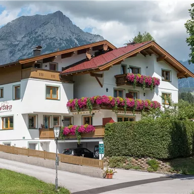 Ein charmantes Haus mit bunten Blumen auf den Balkonen. Im Hintergrund sind majestätische Berge und ein blauer Himmel zu sehen.