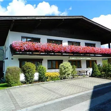 Ein hübsches Haus mit einem blühenden Balkon und einem gepflegten Garten. Im Hintergrund sind Berge und ein blauer Himmel zu sehen.