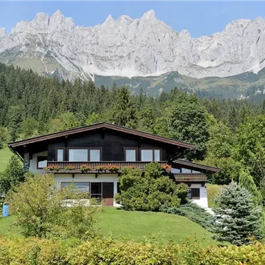 Ein schönes Haus in einer grünen Landschaft mit Bergen im Hintergrund. Die Umgebung ist voller Bäume und Sträucher.