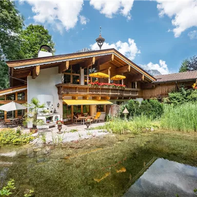 A charming wooden house with a beautiful garden and a small pond. In the background, there are trees and a bright blue sky.