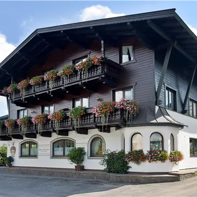 Ein traditionelles alpine Haus mit Holzfassade und blühenden Balkonen. Es liegt umgeben von grünem Gras und hat einen klaren blauen Himmel im Hintergrund.