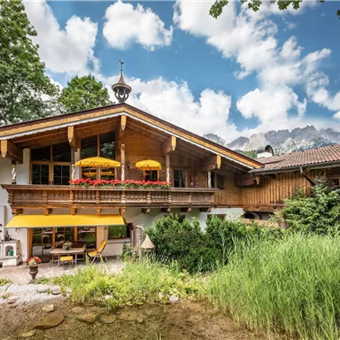 A charming house in alpine style with a large balcony and colorful sunshades. Surrounded by green meadows and a clear sky.