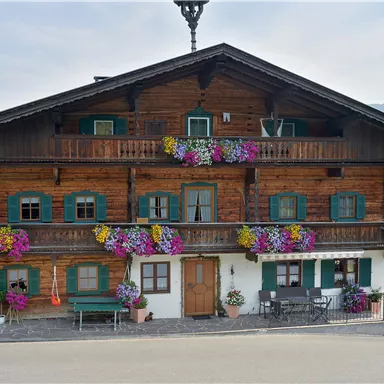 Ein traditionelles Holzhaus mit bunten Blumen an den Balkonen. Im Hintergrund sind sanfte Hügel und ein klarer Himmel zu sehen.