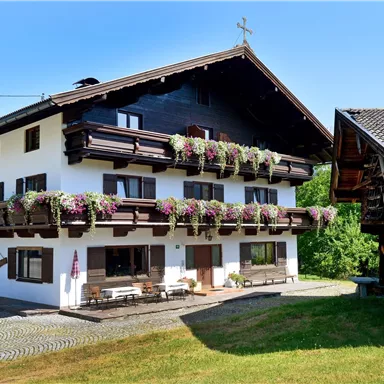 Ein malerisches Haus im alpenländischen Stil mit Balkonen, die mit bunten Blumen geschmückt sind. Der Himmel ist klar und die Umgebung ist grün und einladend.