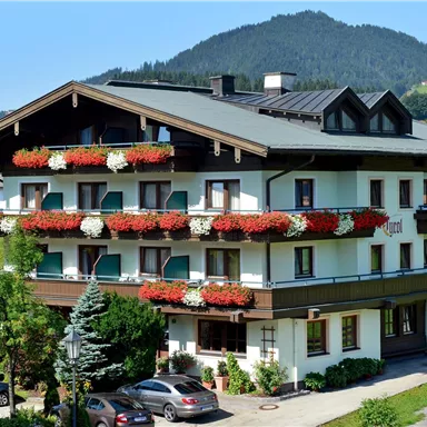 Ein charmantes Hotel mit blühenden Balkonpflanzen und einem malerischen Bergblick. Die Fassade ist freundlich und einladend, umgeben von Grünflächen.