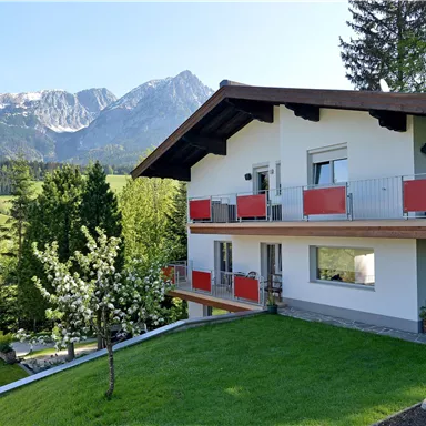 Ein modernes Haus umgeben von Bäumen und einem gepflegten Garten. Im Hintergrund sind beeindruckende Berge und ein klarer Himmel zu sehen.
