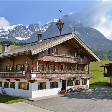 A traditional alpine house surrounded by green meadows and high mountains. Snow-capped peaks are visible in the background.
