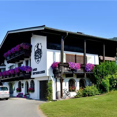 A picturesque building in alpine style with colorful flowers on the balcony. Surrounded by well-kept greenery and a clear blue sky.