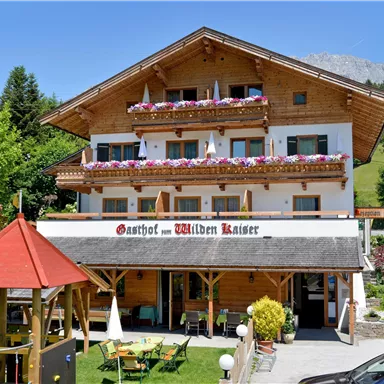 An inviting wooden inn with colorful flowers on the balcony. Surrounded by trees and a green landscape.