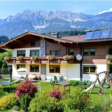 Ein charmantes Haus mit Blumenbalkonen und eine schöne Gartenlandschaft. Im Hintergrund sind majestätische Berge zu sehen.