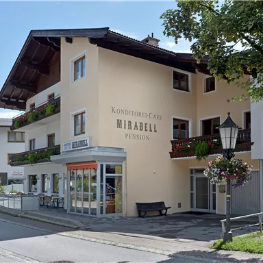 A charming guesthouse decorated with balconies and flowers. The facade is bright and inviting, surrounded by trees and a peaceful street scene.