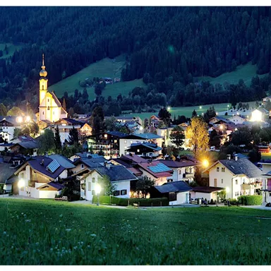 Ein malerisches Dorf bei Nacht, umgeben von sanften Hügeln. Die Kirche mit ihrem beleuchteten Turm ist ein Blickfang in der idyllischen Landschaft.