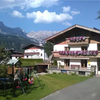Ein charmantes Haus mit blühenden Balkonblumen und einem gepflegten Garten. Im Hintergrund sind majestätische Berge und ein blauer Himmel zu sehen.