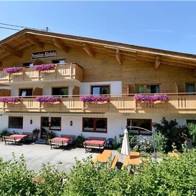 A Alpine guesthouse with a wooden balcony adorned with carvings and colorful flower boxes. The surroundings are green and idyllic in the sunny light.