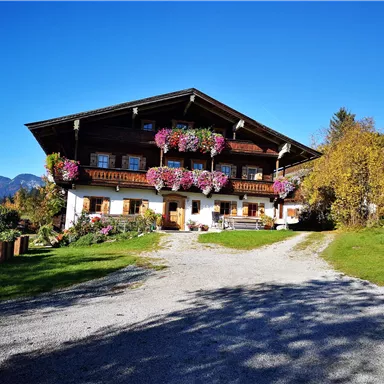A traditional chalet with colorful flowers on the balcony. The sky is clear and blue, surrounded by green meadows and mountains in the background.