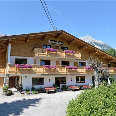 A charming wooden building with balconies, adorned with flowers. In the background, mountains and a blue sky are visible.