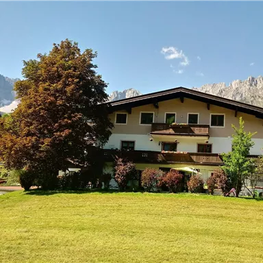 A picturesque building surrounded by a green garden and trees. In the background, majestic mountains can be seen.