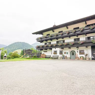 Ein traditionelles Alpenhaus mit mehreren Balkonen und blühenden Blumenkästen. Im Hintergrund sind sanfte Berge und eine grüne Wiese zu sehen.