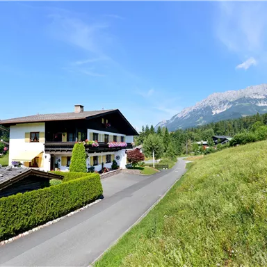 Ein charmantes Haus in einer ländlichen Umgebung mit grünen Wiesen und Bergen im Hintergrund. Der Himmel ist klar und die Landschaft ist sonnig.
