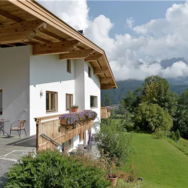 A modern house with a large balcony and colorful flowers. In the background, green meadows and mountains can be seen under a clear sky.