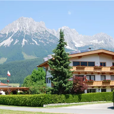 Ein charmantes Haus in den Bergen mit einer grünen Wiese und hohen Bäumen. Im Hintergrund erheben sich majestätische Berge unter einem klaren blauen Himmel.