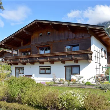 Ein traditionelles Almhütte mit Holzverkleidung und einem gepflegten Garten. Im Hintergrund sind Bergen und blauer Himmel zu sehen.