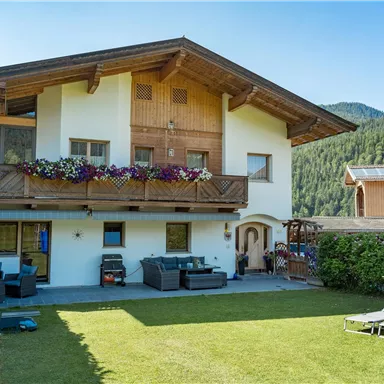 Ein gemütliches Haus mit einem Garten in den Bergen. Das Gebäude hat einen Balkon mit Blumen und ist von grünen Wiesen umgeben.