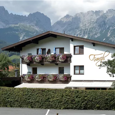 A picturesque house with balconies full of flowers in front of an impressive mountain backdrop. The landscape is green and inviting for relaxation.