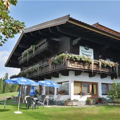 A charming Alpine house with balconies full of flowers. The well-maintained garden has tables and chairs, as well as two blue umbrellas.