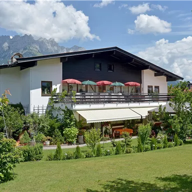 Ein modernes Haus mit einem gepflegten Garten und Blick auf die Berge. Der Himmel ist blau mit einigen Wolken und es gibt Sonnenschirme auf der Terrasse.