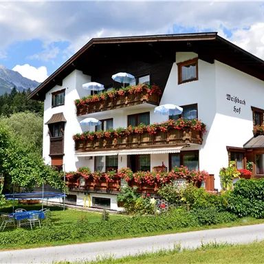 A charming building with flower boxes and white sun umbrellas. Surrounded by a green meadow and mountains in the background.